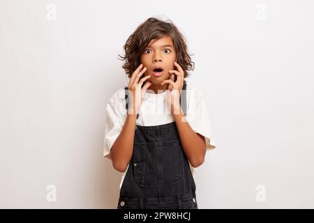 Ein verblüffter junger, dunkelhäutiger Kerl mit lockigem Haar, angezogen in Overalls und einem weißen T-Shirt. Hochwertiges Foto Stockfoto