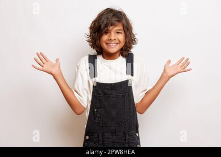 Fröhlicher brauner Teenager mit lockigem Haar, lächelt zahm, verteilt Handflächen, trägt lässige Kleidung, posiert auf weißem Hintergrund. Hochwertiges Foto Stockfoto