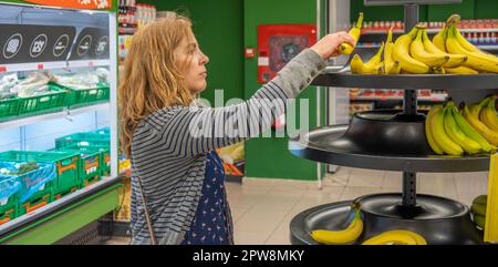 Reife, hübsche blonde Frau, die sich Sorgen machte und über die hohen Preise erstaunt, als sie Bananen aus einem Regal kaufte, während sie Lebensmittel in einem Supermarkt einkaufte. Stockfoto