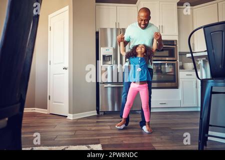 Wie bist du so groß geworden, Dad? Ein glückliches kleines Mädchen, das auf den Füßen ihres Vaters balanciert, während sie zusammen in der Küche herumlaufen. Stockfoto