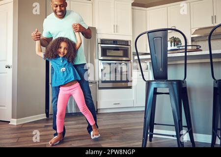 Sie unterhalten sich gegenseitig. Ein glückliches kleines Mädchen, das auf den Füßen ihres Vaters balanciert, während sie zusammen in der Küche herumlaufen. Stockfoto
