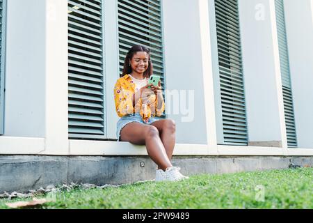 afroamerikanische Studentin, die mit dem Handy lächelt, tolle Nachrichten liest oder online einkauft und ihre Freizeit auf dem Universitätscampus genießt. Glückliche Teenagerin, die auf das Handy schaut. Hochwertiges Foto Stockfoto