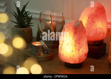 Salzlampen, Kerzen, Zimmerpflanzen und Edelsteine auf einem Holztisch in der Nähe weißer Innenwände. Bokeh-Effekt Stockfoto