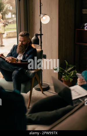 Ein gutaussehender bärtiger Mann, der etwas auf seinen Notizblock schreibt, mit überkreuzten Beinen, der bequem auf dem Stuhl im Café sitzt Stockfoto