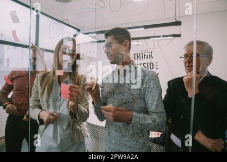 Nahaufnahme von Geschäftsleuten, die eine Diskussion führen, während sie Haftnotizen auf das Glas legen, während sie im CO-Arbeitsbereich stehen Stockfoto