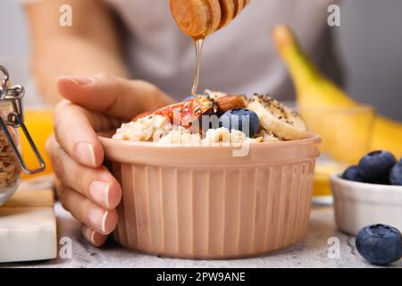 Frau, die Honig zu Haferflocken mit Heidelbeeren, Mandeln, Bananen und Feigenstücken am weißen Tisch hinzufügt, Nahaufnahme Stockfoto