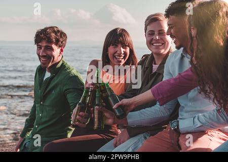 Eine vielfältige Gruppe von Freunden, die mit Bierflaschen anstoßen, während sie an einem felsigen Ufer am Meer sitzen. Stockfoto