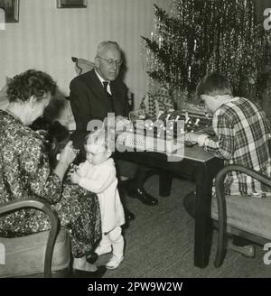 Deutschland. 1960er. Großeltern mit ihren Enkelkindern zu Weihnachten. Ein Großvater spielt mit seiner Enkelin ein Tischfußballspiel, während eine Großmutter auf ihr kleines Enkelkind achtet. Im Hintergrund befindet sich ein Weihnachtsbaum mit Lametta Lametta Lametta Lametta Lametta und Kerzen. Stockfoto