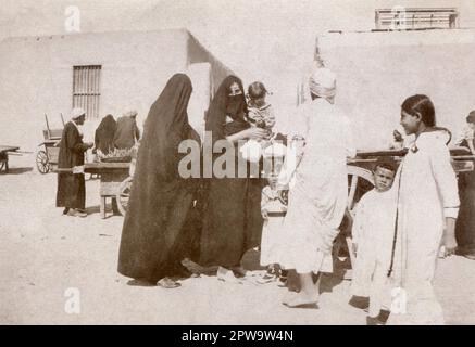 Ägypten. Etwa 1918. Frauen mit ihren Kindern kaufen auf einem Markt ein. Die Frauen tragen al-habara, bestehend aus einem langen Rock, einem Kopfbezug und al-burqu“, einem Gesichtsbezug. Eine Frau trägt auch Arūsat al Burqa, Ornamente auf dem Gesichtsschleier. Der Straßenverkäufer trägt eine Gallibaya und ein Armband, die zeigen, dass er ein offiziell anerkannter „carter“ ist. Stockfoto