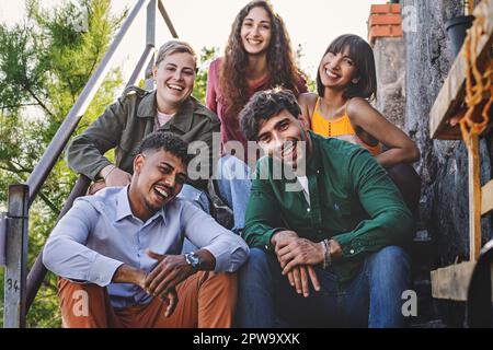 Eine Gruppe von fünf verschiedenen Freunden, die bei Sonnenuntergang auf einer Treppe in einem Touristenort posieren. Sie lächeln, schauen in die Kamera und genießen ihren tim Stockfoto