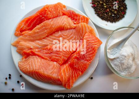 Fischfilet aus frischem rohem Lachs auf weißem Teller, eine kleine Schüssel Salz und schwarze Pfefferkörner auf weißem Hintergrund. Gesundes Essen. Stockfoto