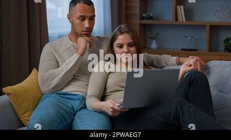 Multiethnisches Paar Hausbesitzer, die online einkaufen, verwenden Laptop-Umarmung auf dem Sofa zu Hause vielfältige Frau Frau Frau Frau Frau und Mann Ehemann reden über die Wohnung in echt wählen Stockfoto