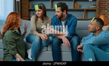 Eine Gruppe von multiethnischen Freunden spielt zu Hause ein Scharadadenspiel, das fröhlich lacht, verschiedene ethnische Männer und Frauen, die raten, wie eine Person mit Haftnotizen spielt Stockfoto