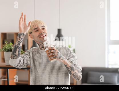 Junger tätowierter Mann mit einer Tasse Kaffee, der zu Hause mit der Hand winkt Stockfoto