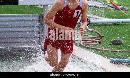 Vorderansicht eines jungen Highschool-Läufers, der während eines Rennens im Wasser der Steeplechase planscht. Stockfoto