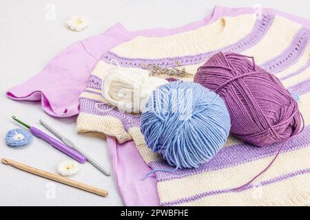 Handgestricktes Baby-T-Shirt in Fliedertönen. Sachen und Requisiten enthalten Fäden, Haken, Stricknadeln und handgefertigte Dekoration. Heller Steinbeton, Backgro Stockfoto