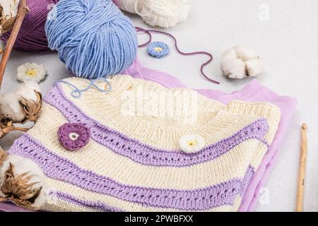 Handgestricktes Baby-T-Shirt in Fliedertönen. Sachen und Requisiten enthalten Fäden, Haken, Stricknadeln und handgefertigte Dekoration. Heller Steinbeton, Backgro Stockfoto