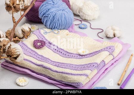 Handgestricktes Baby-T-Shirt in Fliedertönen. Sachen und Requisiten enthalten Fäden, Haken, Stricknadeln und handgefertigte Dekoration. Heller Steinbeton, Backgro Stockfoto