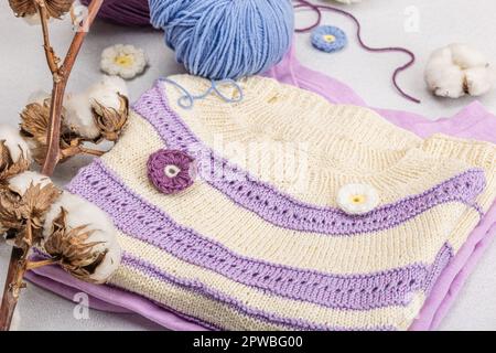 Handgestricktes Baby-T-Shirt in Fliedertönen. Sachen und Requisiten enthalten Fäden, Haken, Stricknadeln und handgefertigte Dekoration. Heller Steinbeton, Backgro Stockfoto
