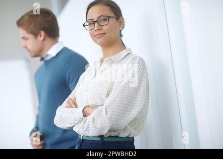 Erfolg in Schritt. Porträt einer attraktiven jungen Frau im Büro mit einem Kollegen im Hintergrund. Stockfoto