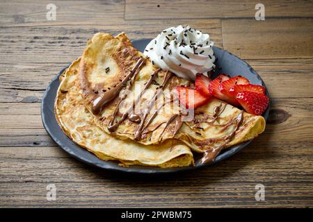 Teller mit Crepes, dekoriert mit Schlagsahne und Erdbeeren auf einem Holztisch Stockfoto