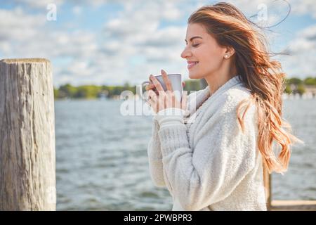 Der Morgen ist geplatzt und der Kaffee hat gesprochen. Eine wunderschöne junge Frau, die ein warmes Getränk auf einem Pier am See genießt. Stockfoto