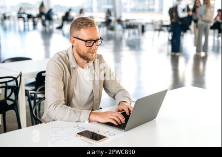 Foto eines fokussierten erfolgreichen weißen Mannes mit Brille und lässiger, eleganter Kleidung, IT-Spezialist, Entwickler, Programmierer, der in einem Laptop in einem Co-working Center arbeitet und ein neues Programm entwickelt Stockfoto