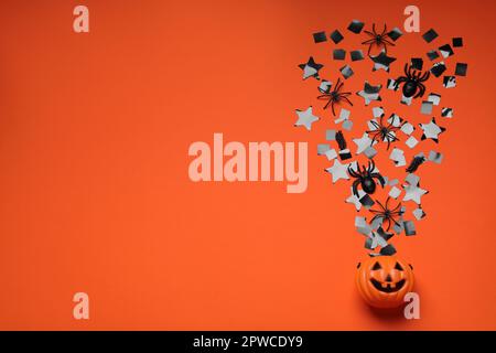 Kürbiskorb aus Kunststoff, schwarzes Konfetti, Spinnen und Platz für Text auf orangefarbenem Hintergrund, flach liegend. Halloween-Feier Stockfoto