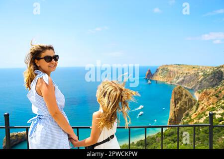 Mutter und kleines Mädchen stehen neben dem Zaun auf Aussichtspunkt und bewundern den malerischen Blick auf die felsige Küste an sonnigen Tagen mit klarem Himmel und blauem Wasser Stockfoto