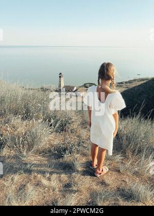 Ganzkörper-Rückansicht von einsamen unkenntlich Mädchen in weiß Sommerkleid steht auf grasbewachsenen Hügel an der Küste mit Leuchtturm Turm Stockfoto