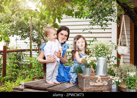 Junge, entzückte Mutter sitzt im Sessel mit entzückenden kleinen Geschwistern auf der Terrasse des Landhauses und entspannt sich am Wochenende zusammen Stockfoto