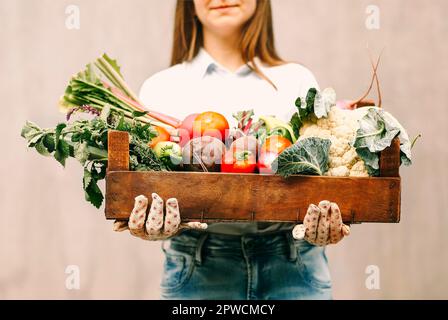 Nicht erkennbare Frau in Handschuhen, die eine Holzkiste mit verschiedenen Gemüsesorten vorführt Während der Arbeit auf dem Bauernhof Stockfoto