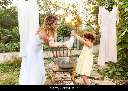 Charming kleines Mädchen helfen Mutter mit Weidenkorb während der Arbeit Chore und hängende Wäsche im Hinterhof im Sommer Abend Stockfoto