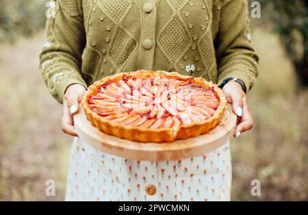 Crop weiblich in Kleid und Strickjacke trägt Brett mit leckeren Kuchen auf verschwommenem Hintergrund der Natur Stockfoto