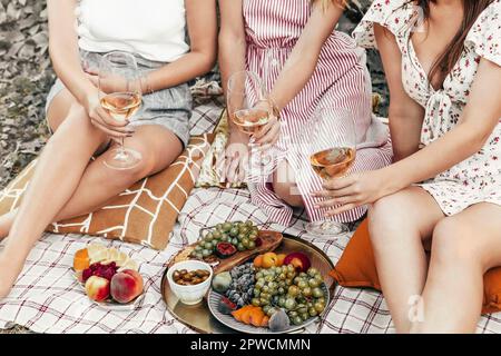 Von oben anonymen Freundinnen klirren Gläser Wein, während sie auf Decken neben Tellern mit Früchten während des Picknicks sitzen Stockfoto