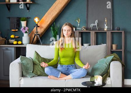 Entspanntes, friedliches, blondes Mädchen der Millennials mit geschlossenen Augen, das im Lotus sitzt, auf der Couch im Wohnzimmer zu Hause posiert und eine Pause für Meditations-Yoga macht Stockfoto