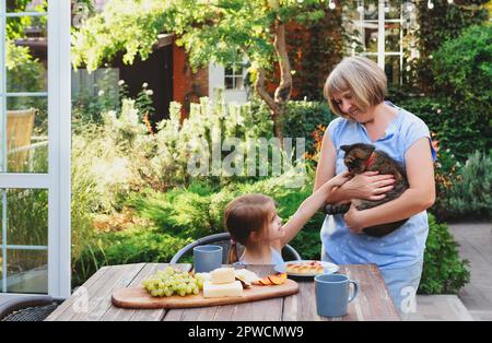 Süßes kleines Mädchen, das am Tisch sitzt und im Garten des Landhauses frühstückt, Katze streichelt, die in den Armen der Großmütter sitzt, Enkeltochter eines Kindes Stockfoto