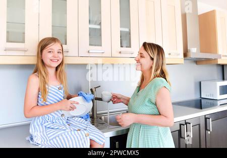 Glückliche mutter und kleine Tochter verbringen gern Zeit damit, bei Hausarbeiten zu helfen, trockenes Geschirr zu waschen, anstatt fröhlich beste Gesellschaft in Sommerkleidern zu tragen Stockfoto