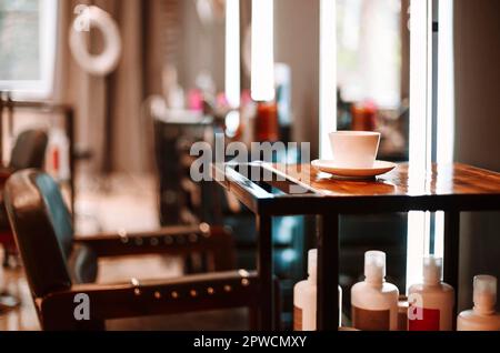Friseursalon im Innenbereich. Kaffeetasse auf Holztisch und Stuhl für Kunden im Kosmetiksalon mit niemandem, beleuchteter Spiegel mit verschwommenem Hintergrund Stockfoto