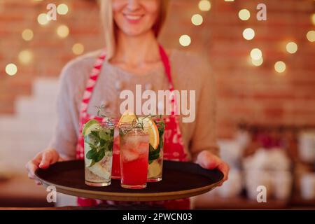 Lächelnde Kellnerin in einer farbigen Schürze mit einem Tablett mit alkoholischen Getränken und verschiedenen Cocktails mit Obstgarnierung in langen Gläsern bei einer festlichen Veranstaltung Stockfoto