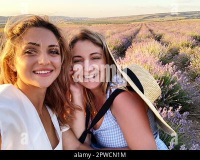 Zwei fröhliche, überfreuliche weibliche Freundinnen, die ein Selfie machen, während sie an einem sonnigen Sommertag im Lavendelfeld stehen und lächelnde Freundinnen machen Stockfoto