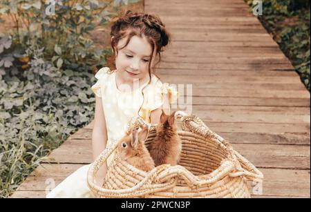 Kleines hübsches Mädchen im Sommerkleid, das draußen auf einem Holzweg vor dem Strohkorb mit Kaninchen sitzt, niedliches Kind, das mit einem leichten Lächeln Hasen ansieht Stockfoto