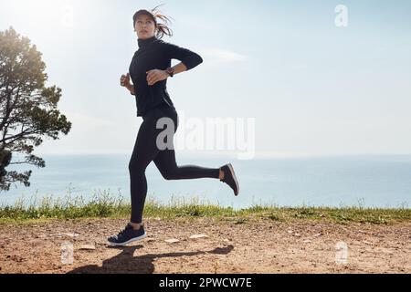 Ich habe noch ein bisschen Abstand. Eine entschlossene junge Frau, die alleine joggen geht und im Hintergrund einen Blick auf das Meer hat. Stockfoto