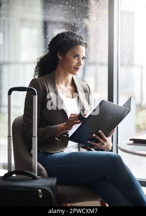 Ein Go Getter zu sein bedeutet, immer in Bewegung zu sein. Eine junge Geschäftsfrau, die in einer Geschäftsmappe liest, während sie auf ihren Flug am Flughafen wartet Stockfoto