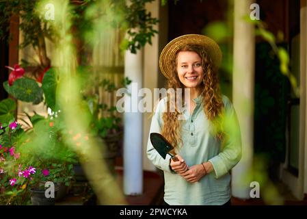 Eine junge Frau steht im Garten und hält Gartenwerkzeug in der Hand. Sie lächelt und schaut in die Kamera. Junge Gärtnerin mit Schaufel in grünem Obstgarten Stockfoto