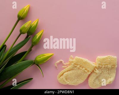 Gelbe Tulpenblumen und gelbe Babyschühchen für ein Neugeborenes auf pinkfarbenem Hintergrund mit Kopierbereich. Muttertag Grußkarte. Wünsche für Mutter schreiben. Knitt Stockfoto