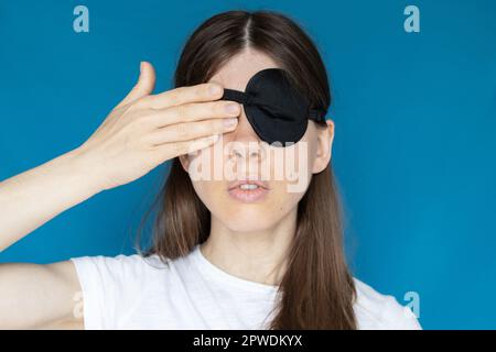 Ein Mädchen mit einem Verband auf dem einen Auge und dem anderen Auge mit der Hand auf blauem Hintergrund, nicht sehen, nicht gucken Stockfoto