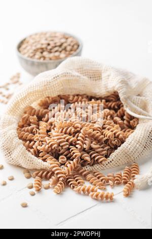 Grüne Fusilli-Nudeln mit Linsen auf weißem Fliesenhintergrund. Beutel mit roher Pasta und Schüssel mit grünen Linsen. Glutenfreie Nudeln. Stockfoto