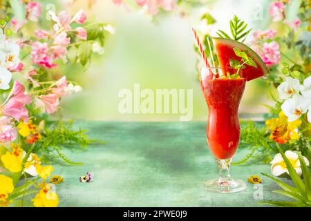 Sommerliche kalte Wassermelone und Erdbeergetränk. Köstlicher Beerencocktail auf einem grünen Tisch, umgeben von tropischen Blättern und Blumen. Stockfoto