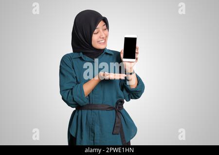 Porträt einer aufgeregten asiatischen muslimischen Frau mit Hijab, die ein Mobiltelefon mit leerem Bildschirm zeigt. Werbemodell-Konzept. Isoliertes Bild auf WHI Stockfoto